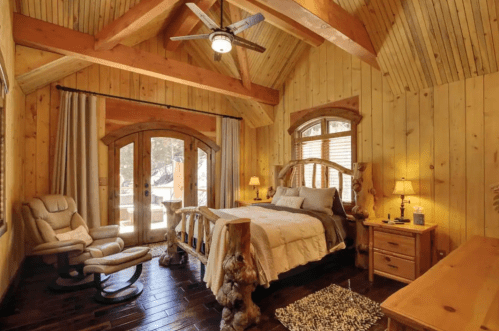 Cozy wooden bedroom with a large bed, rocking chair, and natural light from double doors leading outside.